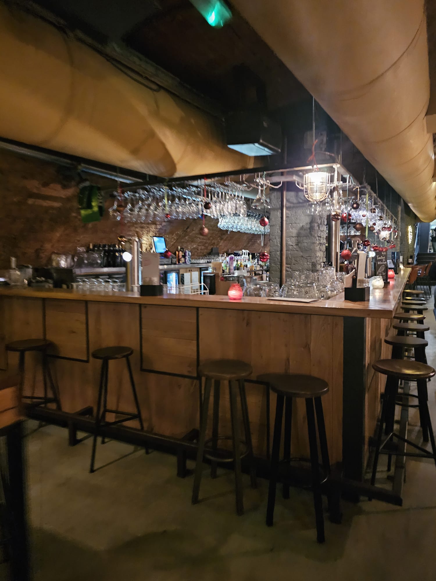 A long wooden bar with black industrial stools under warm, dim lighting