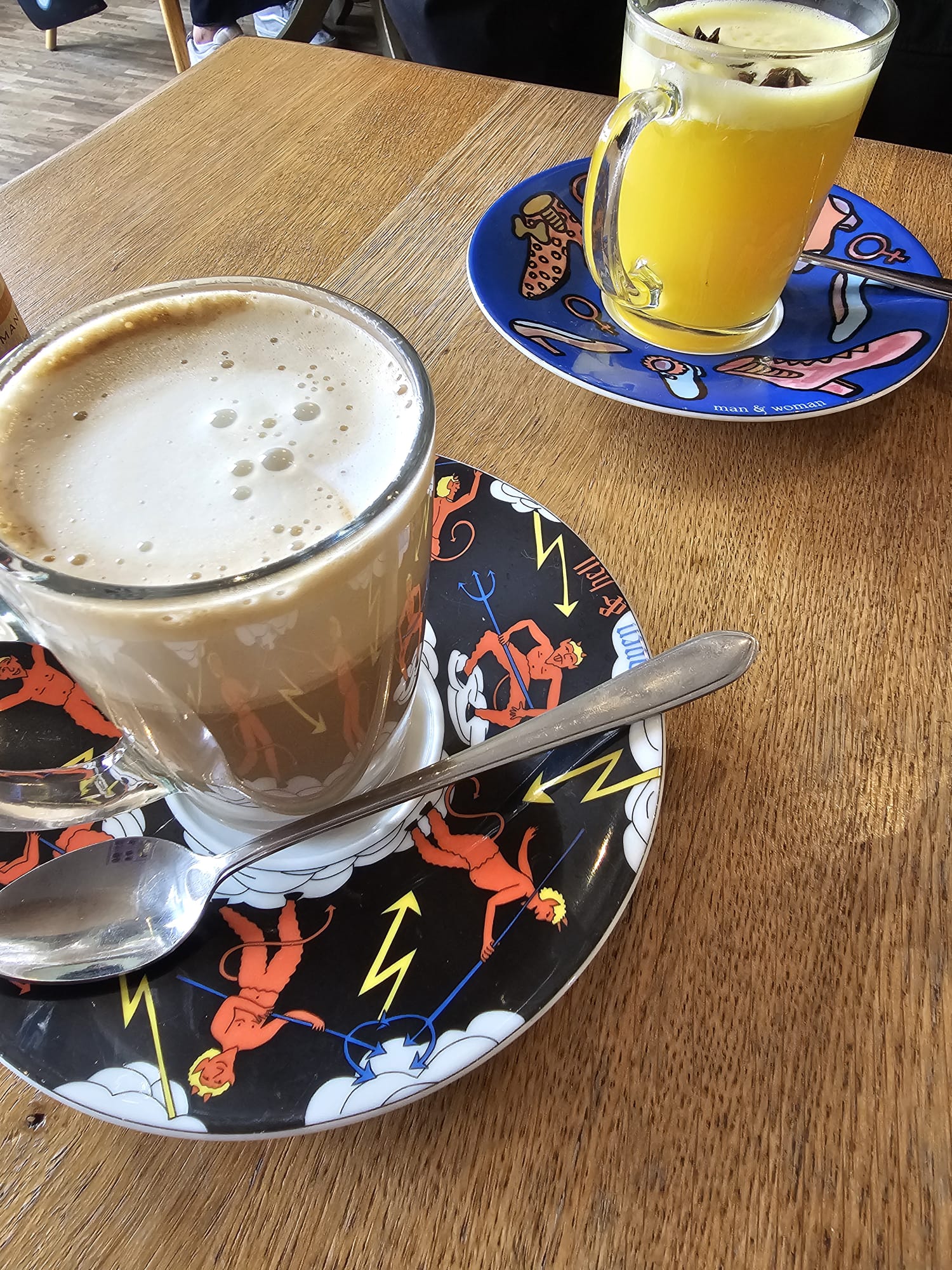 Two unique drinks on a wooden table; a mushroom latte in a glass mug and a vibrant yellow Orange Ginger Dream, both served on colorful, artistic saucers.
