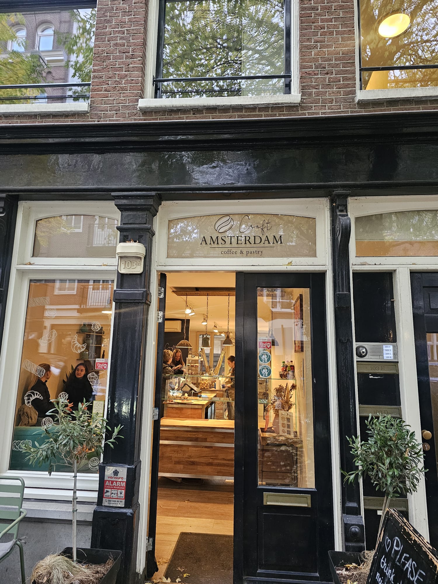 The storefront of Craft Amsterdam, a 100% gluten-free bakery, featuring a classic black and white facade with large glass windows showing a warm, wood-accented interior.