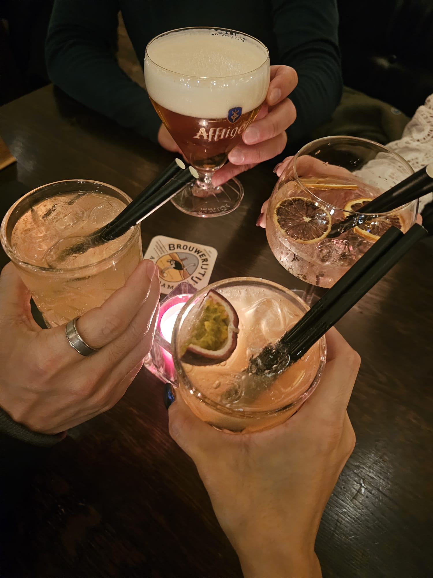 Four people holding up various drinks, including a foamy beer in an Affligem glass and colorful gin and tonics with fruit garnishes.