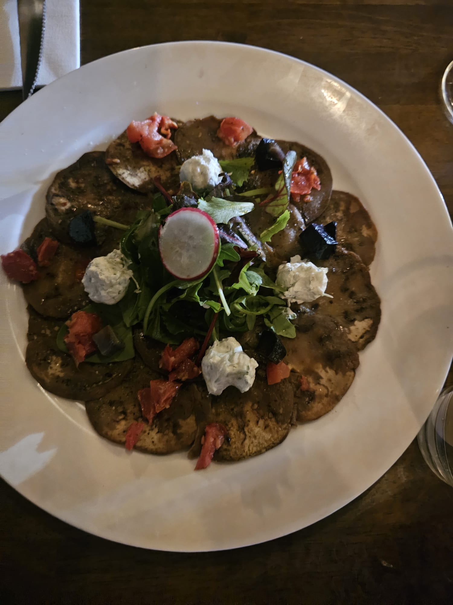 Thinly sliced roasted eggplant arranged in a circle on a white plate, topped with fresh arugula, dollops of white cheese, and radishes.