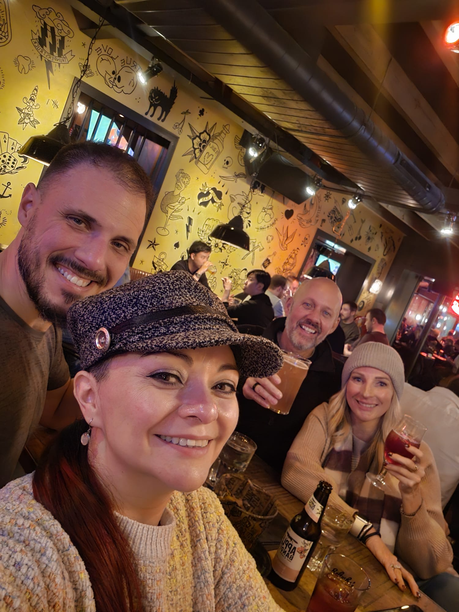 Group of four friends smiling and holding drinks at a lively bar with illustrated yellow walls in Amsterdam
