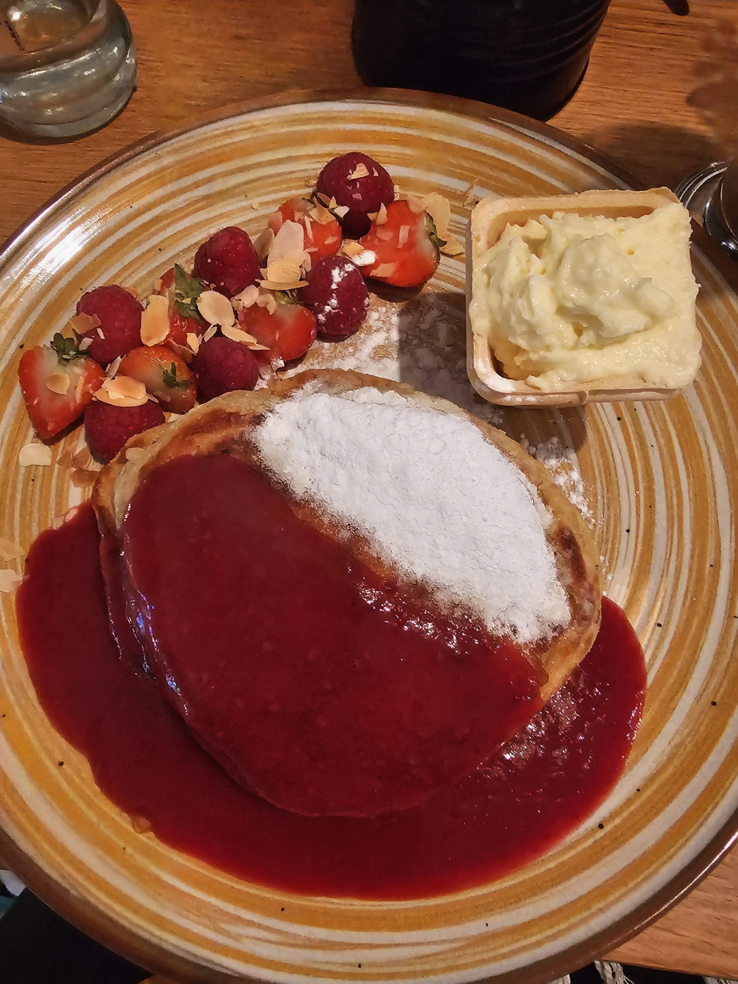 A plate of gluten-free pancakes topped with powdered sugar, fresh strawberries, and a bright red berry coulis, served with a side of whipped cream