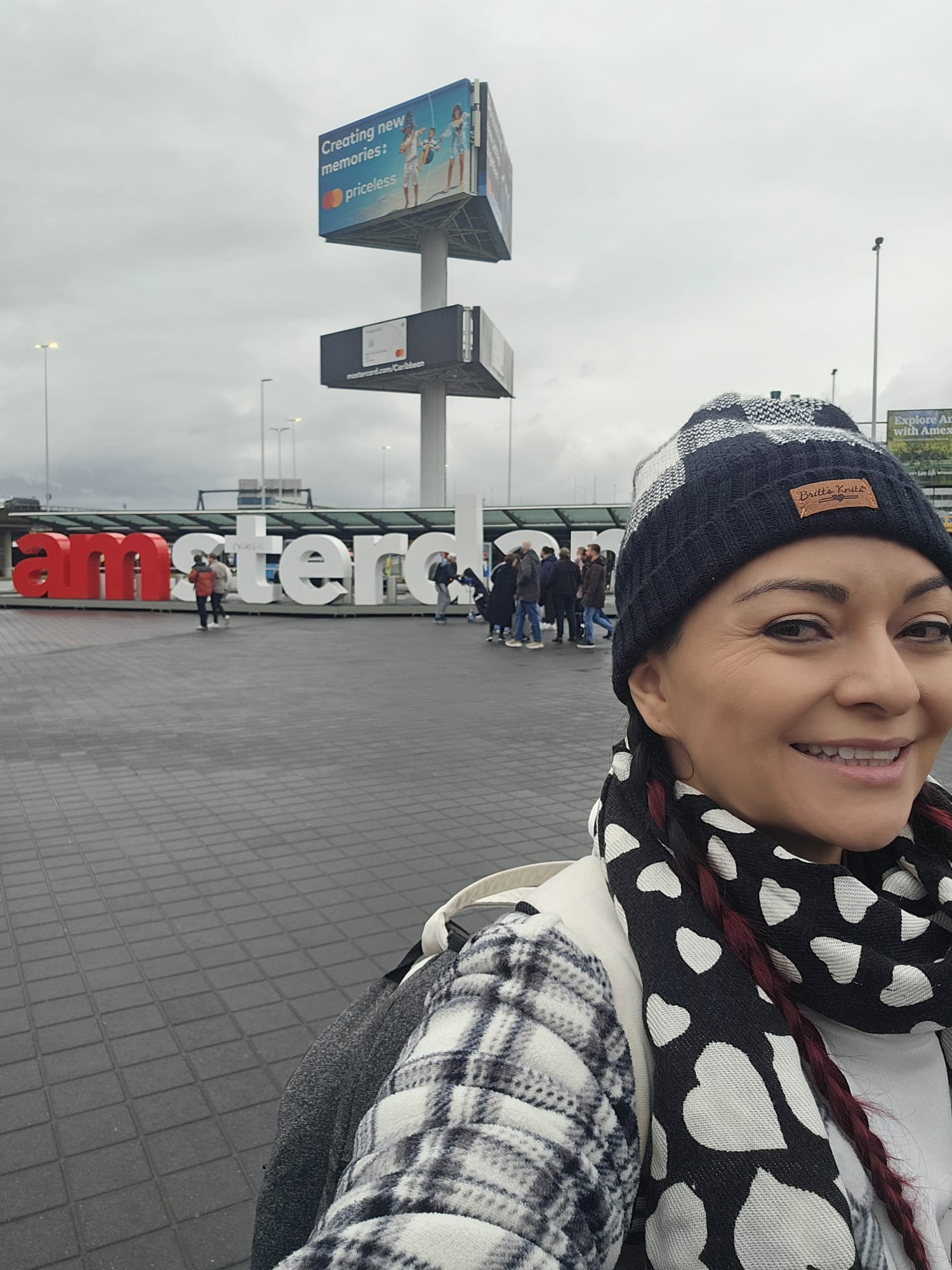 Woman taking a selfie in front of the large Amsterdam sign at Schiphol Airport on a cloudy day, wearing a beanie and heart print scarf