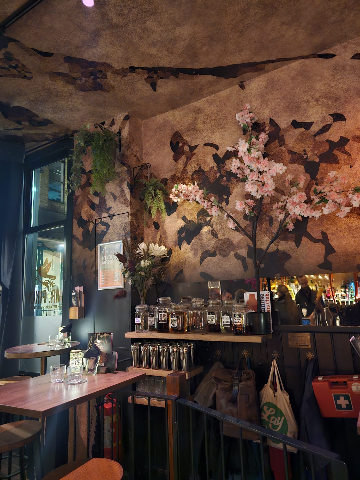 A moody bar interior featuring dark walls and a large floral mural with pink blossoms.
