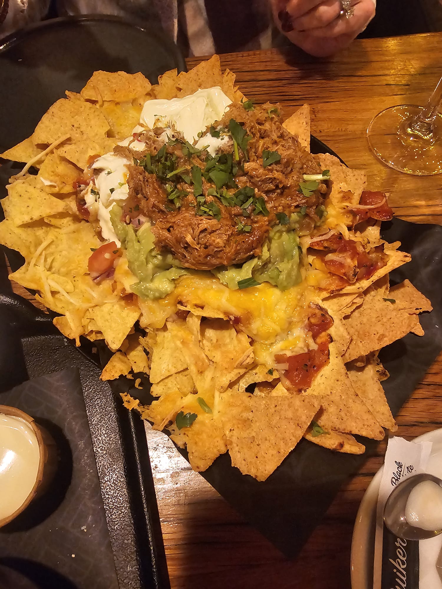 A large platter of tortilla chips piled high with melted cheese, guacamole, sour cream, and tender pulled pork, garnished with fresh cilantro.
