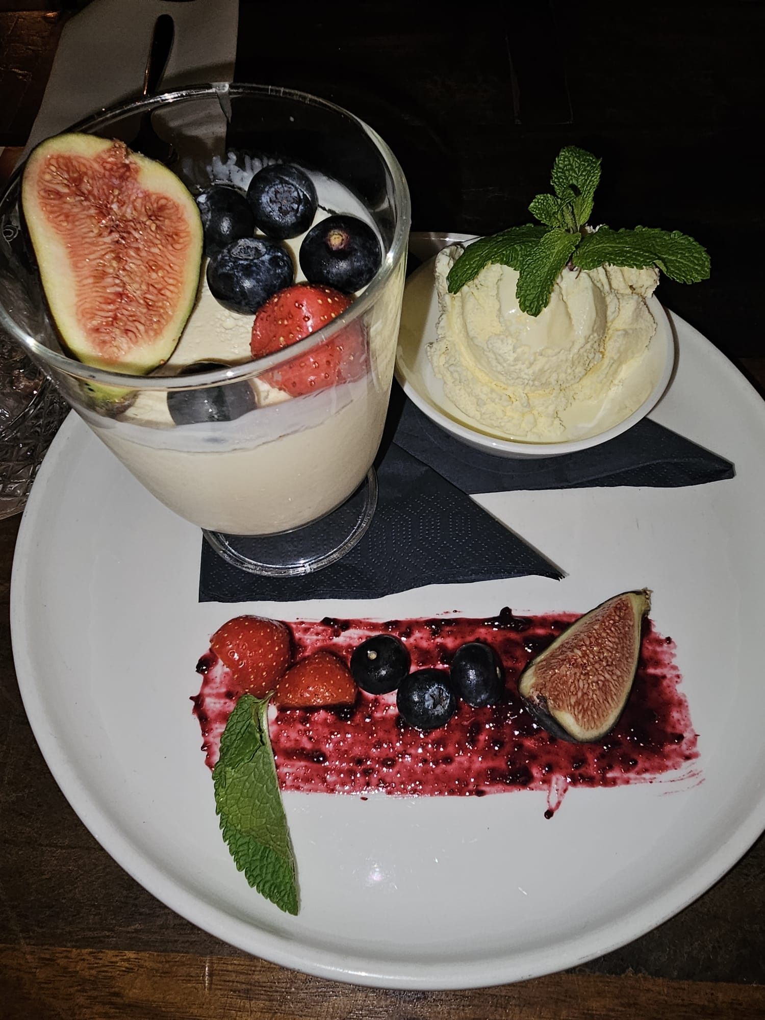 A dessert glass filled with creamy panna cotta, topped with a fresh fig and blueberries, served on a plate with a scoop of vanilla ice cream and a berry smear.