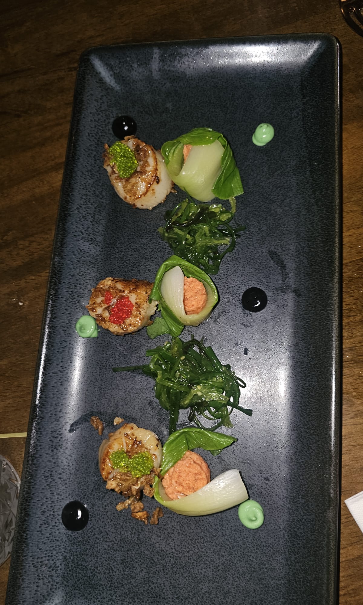 Three seared scallops arranged on a rectangular black plate with seaweed salad and small garnishes of sauce.