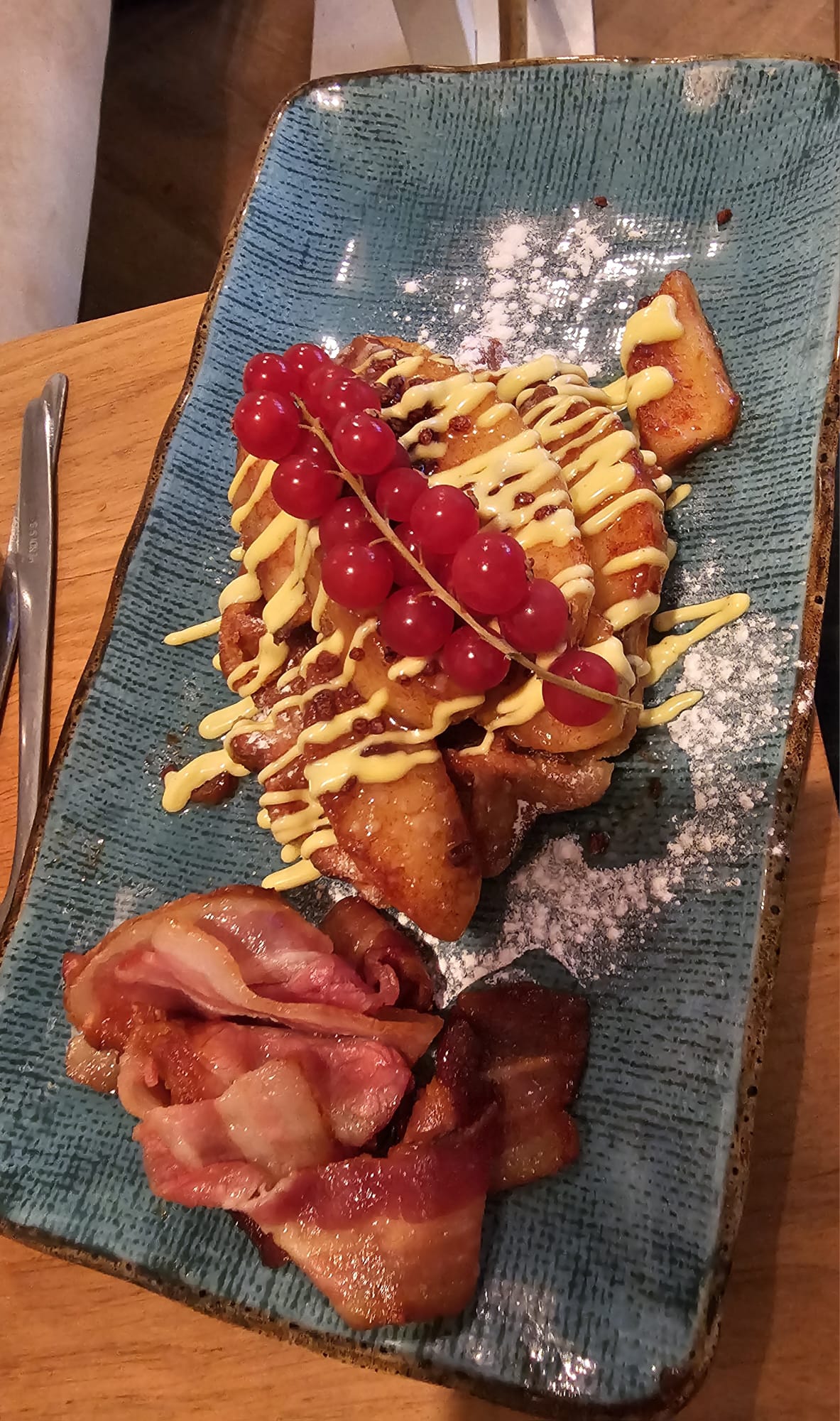 A thick waffle topped with caramelized fruit, a drizzle of yellow custard sauce, and a sprig of red currants, served with a side of crispy bacon.
