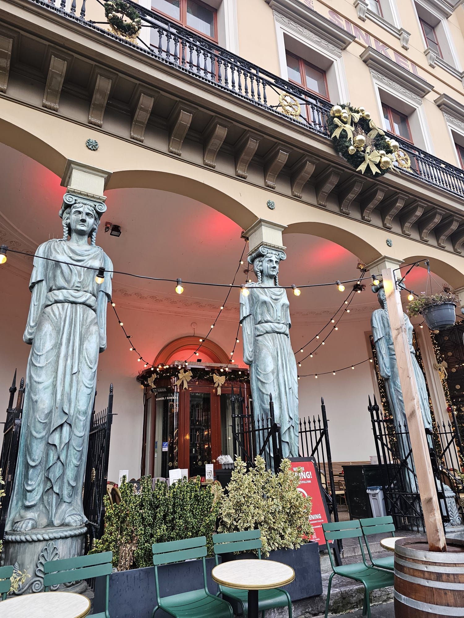 A grand, historic building exterior with stone statues (caryatids) and arched entryways lit with red lights.