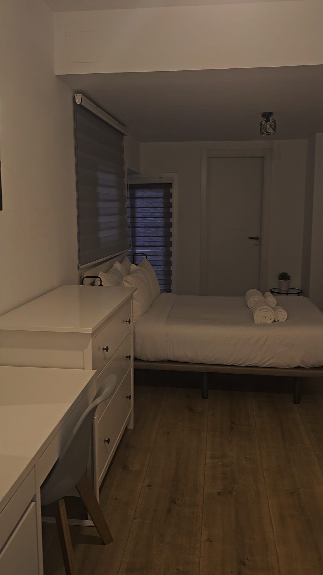 A photo of an ensuite bedroom in a Barcelona apartment, featuring a bed with white linens and rolled-up towels, a white dresser, and a closed door. The room has wooden flooring and is dimly lit