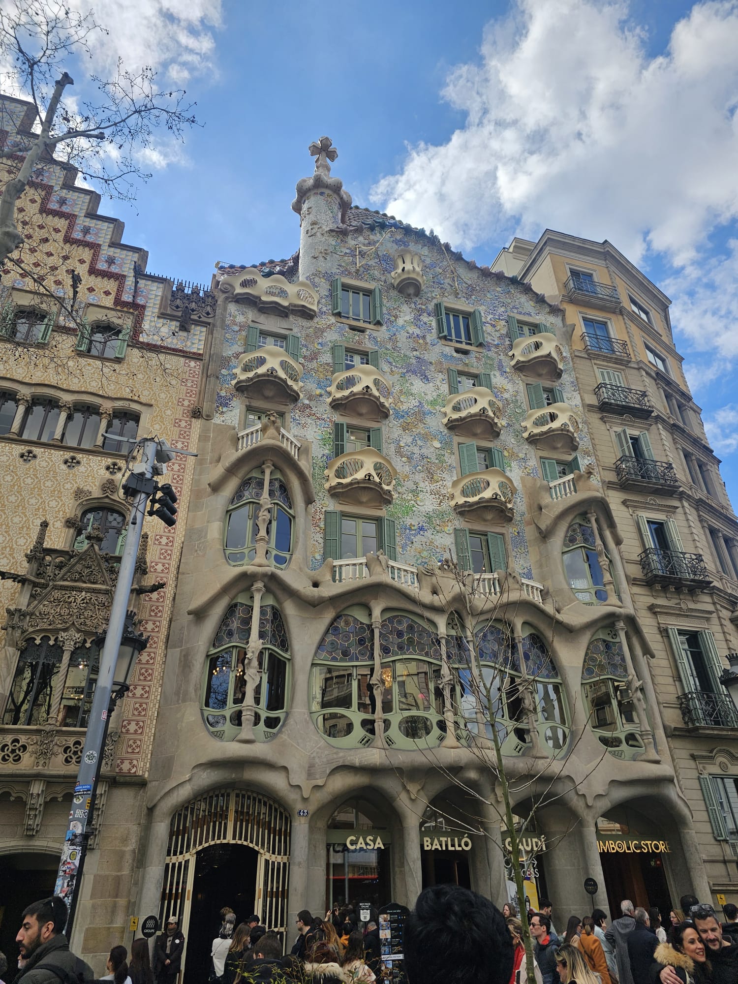 A detailed, low-angle photo of the organic and colorful facade of Casa Batlló in Barcelona, Spain. The building, designed by Antoni Gaudí, features unique elements such as bone-like columns on the ground floor and balconies nicknamed "House of Bones", marine-inspired details, and a vibrant, scaled roofline. 