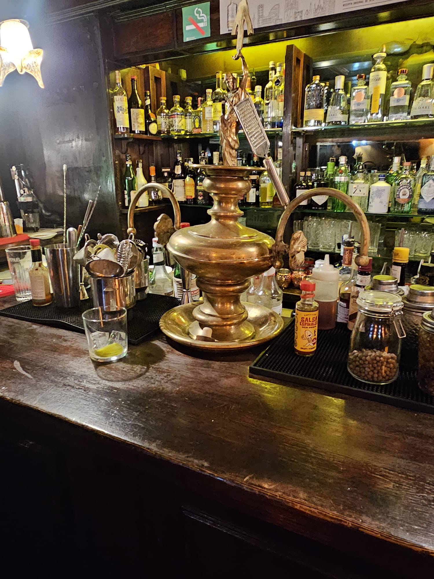 Dark moody bar counter at Cafe Bar l'ascensor featuring an antique brass distillation machine surrounded by bottles of spirits