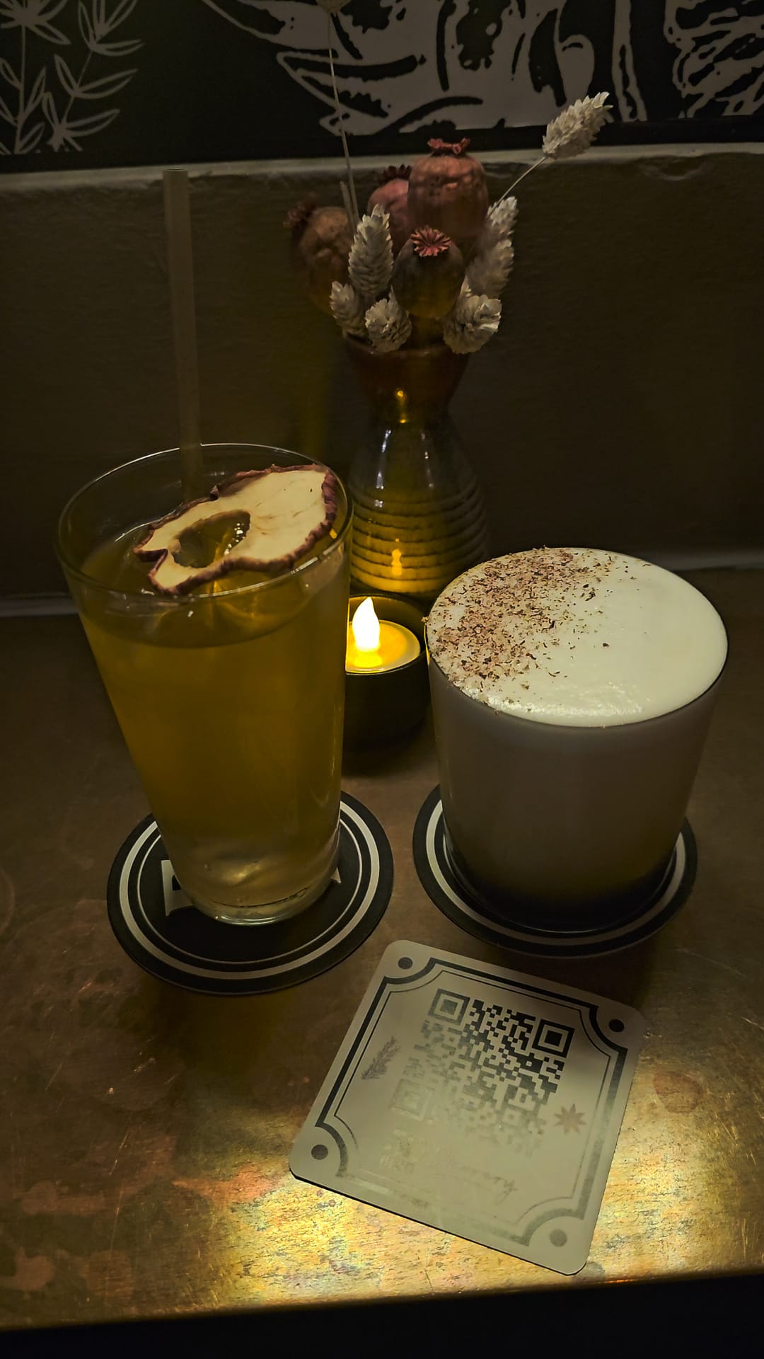 2 cocktails on a table in a dimly lit bar with a vase of flowers behind them