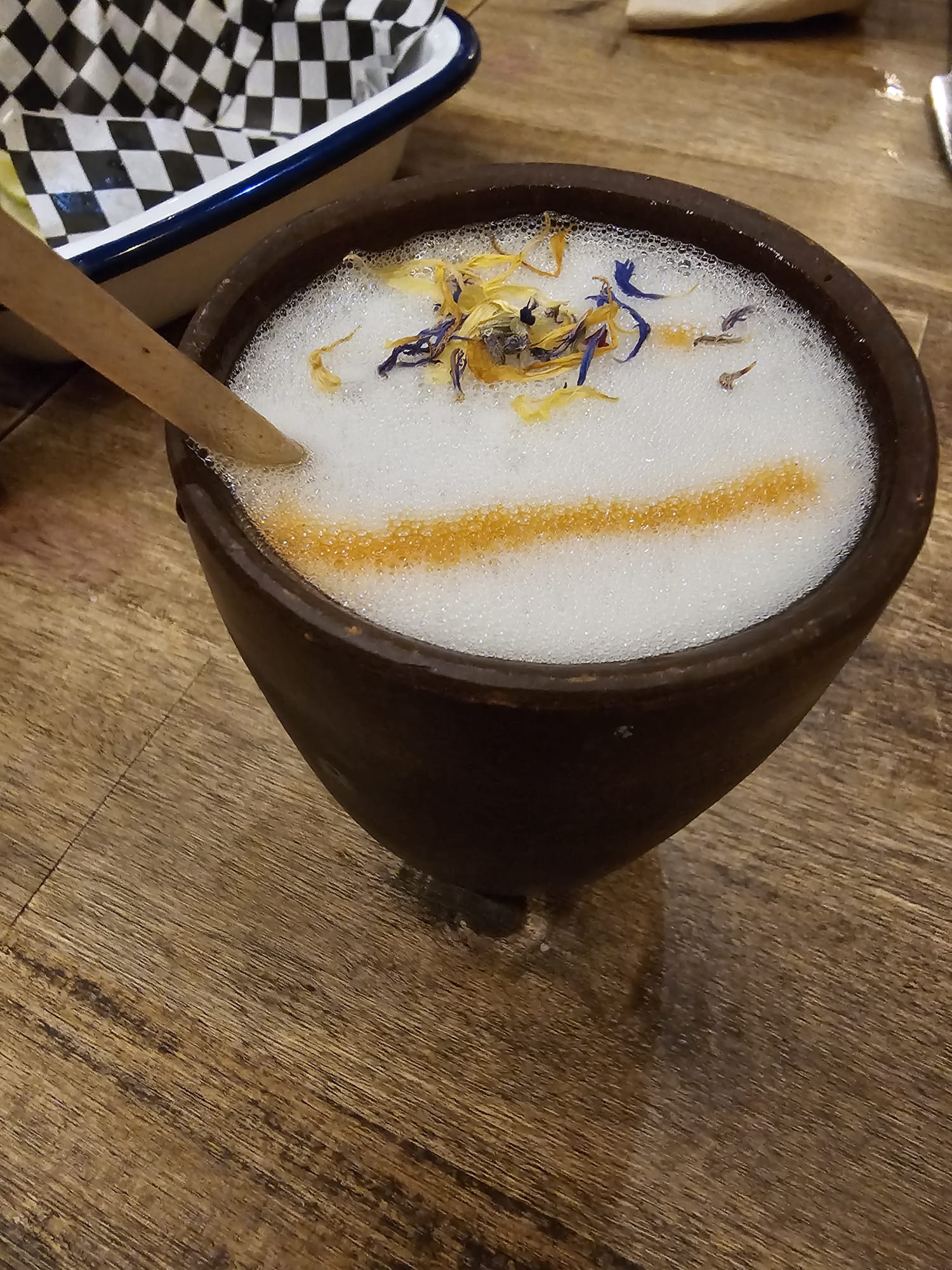 The image shows a cocktail called the Esperanza, presented in a traditional Mexican clay cup (known as a cantarito). The drink is topped with a thick, velvety white foam and garnished with delicate blue and yellow edible flower petals and a distinct line of orange citrus dust or spice. A wooden straw or stirrer rests against the side of the cup