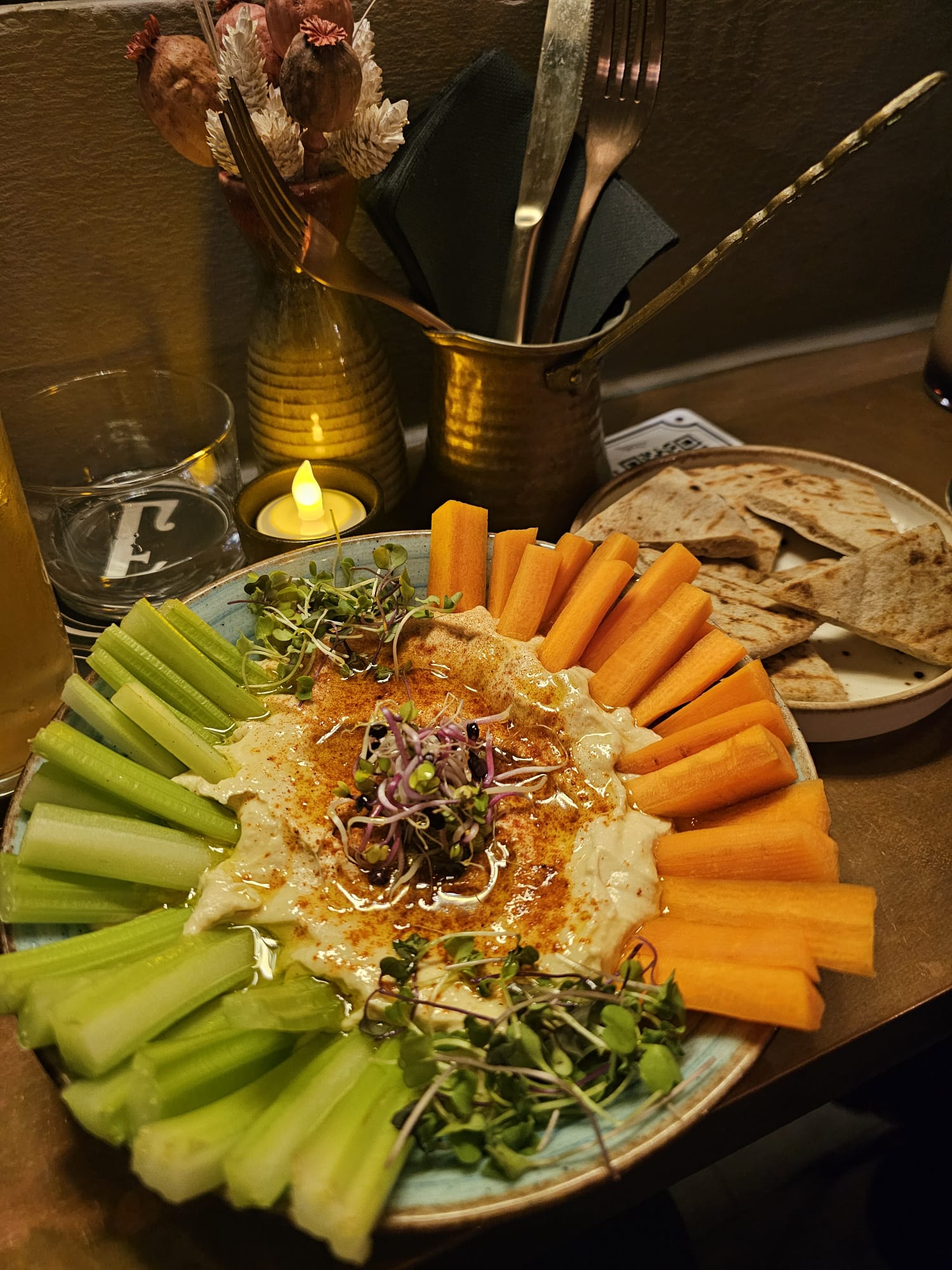Beautifully plated hummus with carrots, cucumber, microgreens, and flatbread crisps with a candle and gold cutlery holder at Especiarium