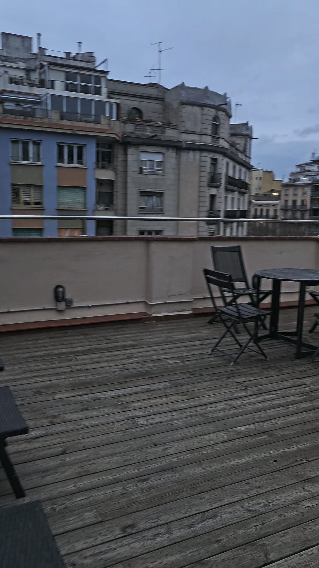 A view from a large wooden-floored apartment balcony in Barcelona, showing outdoor furniture and surrounding city buildings under an overcast sky. Text overlay reads: 'Part of the huge balcony of our Barcelona apartment'."