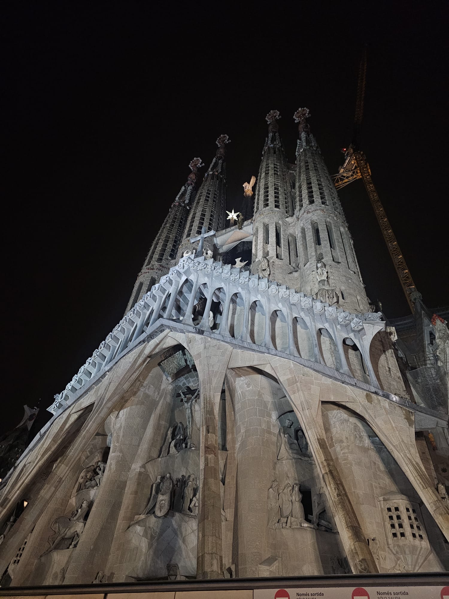 A low-angle, night time photo of the ornate, illuminated exterior of the Sagrada Familia basilica in Barcelona, featuring several tall, spiky spires and scaffolding on one section