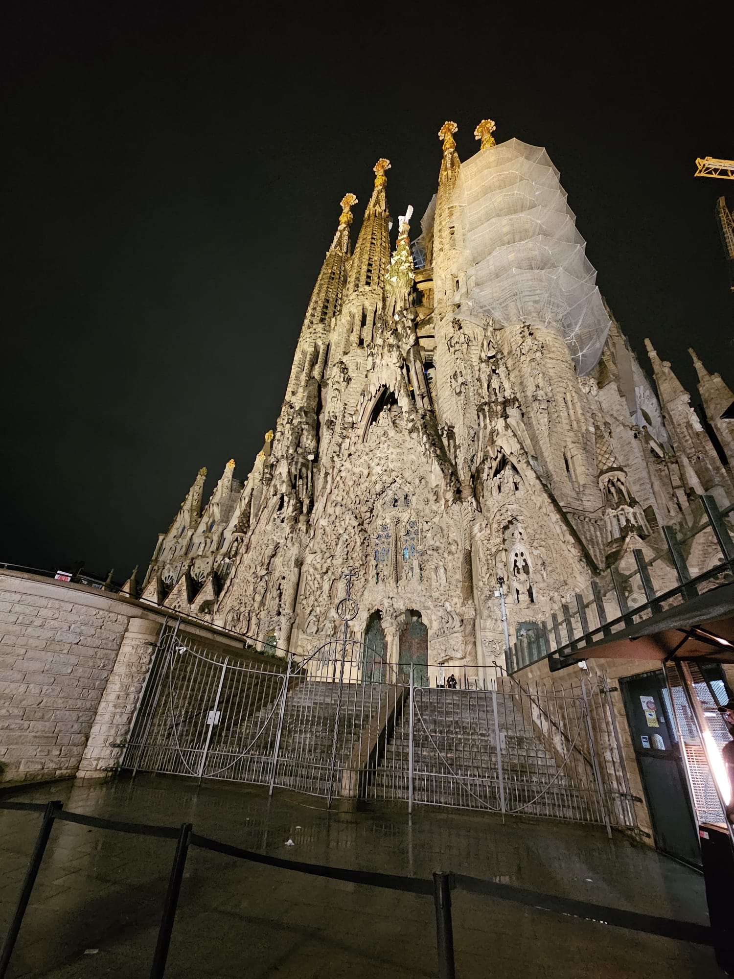 A low-angle photograph of the Sagrada Familia church at night. The intricate stone facade of the Nativity side is brightly illuminated, standing out against the dark sky. Scaffolding is visible on one of the spires on the right side. A metal fence is in the foreground. Text overlay at the bottom reads "La Sagrada Familia at night"