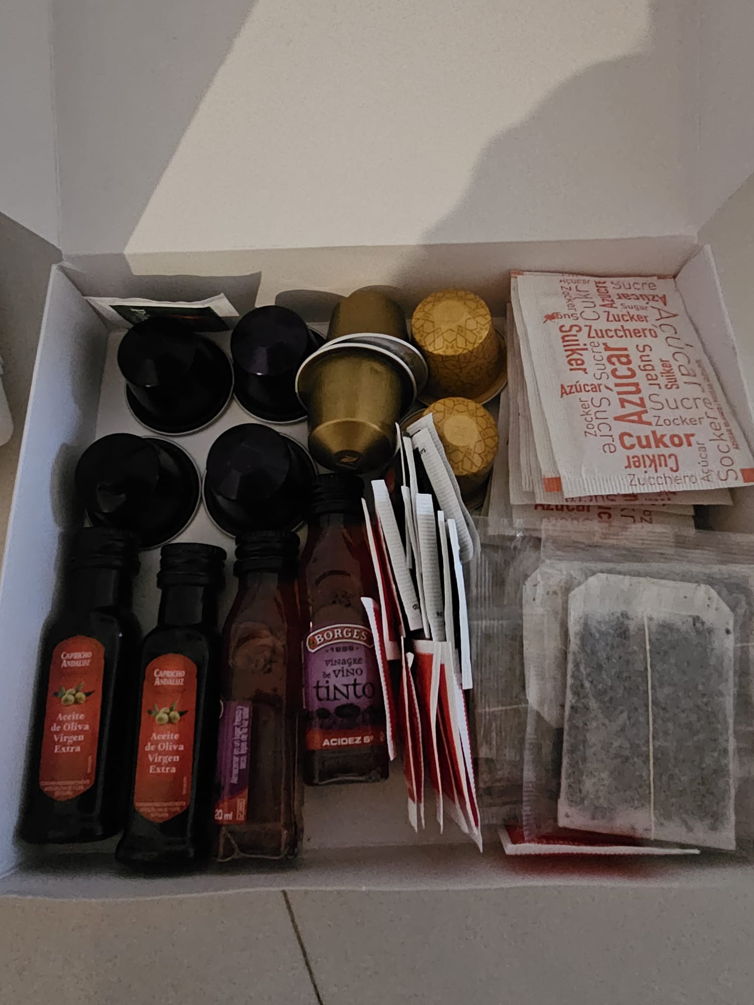 An open box filled with hospitality items, including small bottles of olive oil and red wine, assorted coffee pods, sugar packets in multiple languages, and tea bags. 