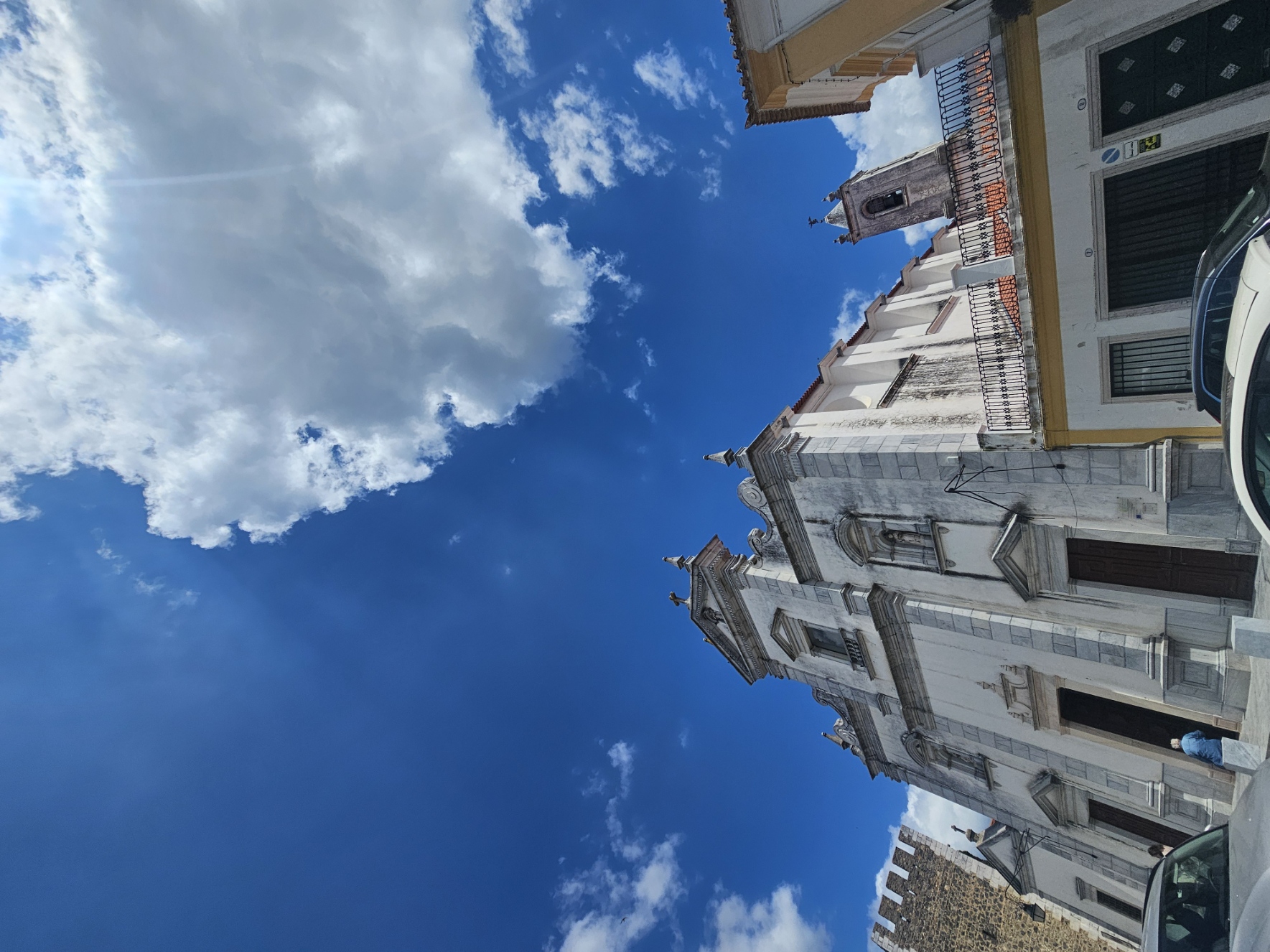 Dramatic upward angle of Beja Cathedral's white baroque facade with twin bell towers reaching into a cloudy blue sky, Portugal