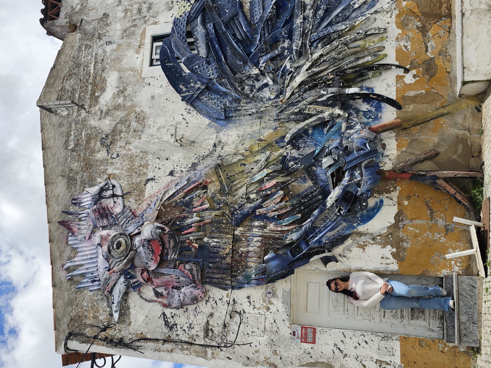 Woman standing in front of a massive street art mural of a rooster made from recycled materials painted on a weathered building wall in Beja, Portugal