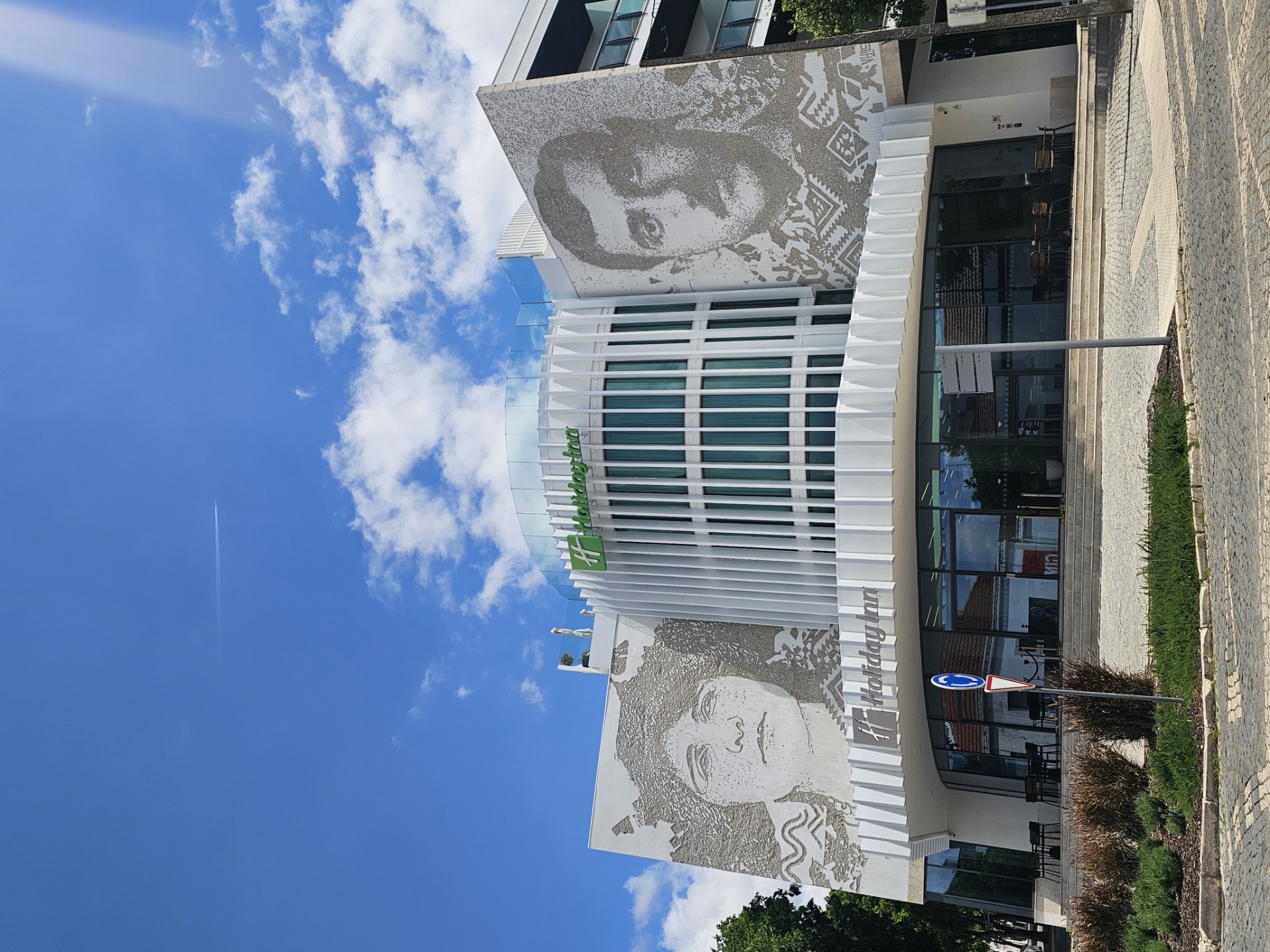 Exterior of the Holiday Inn Beja, a modern white building featuring large-scale portrait murals of two figures on either side of the facade, under a bright blue sky in Beja, Portugal