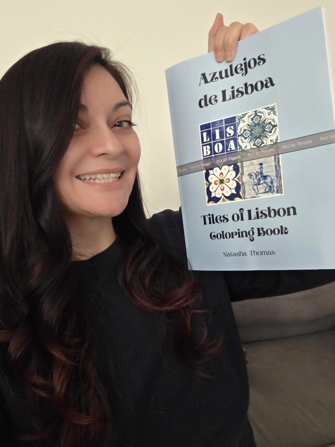 A woman smiling and holding up a coloring book titled "Azulejos de Lisboa: Tiles of Lisbon" against a plain background. The book has a light blue cover with tile designs and the name Natasha Thomas.