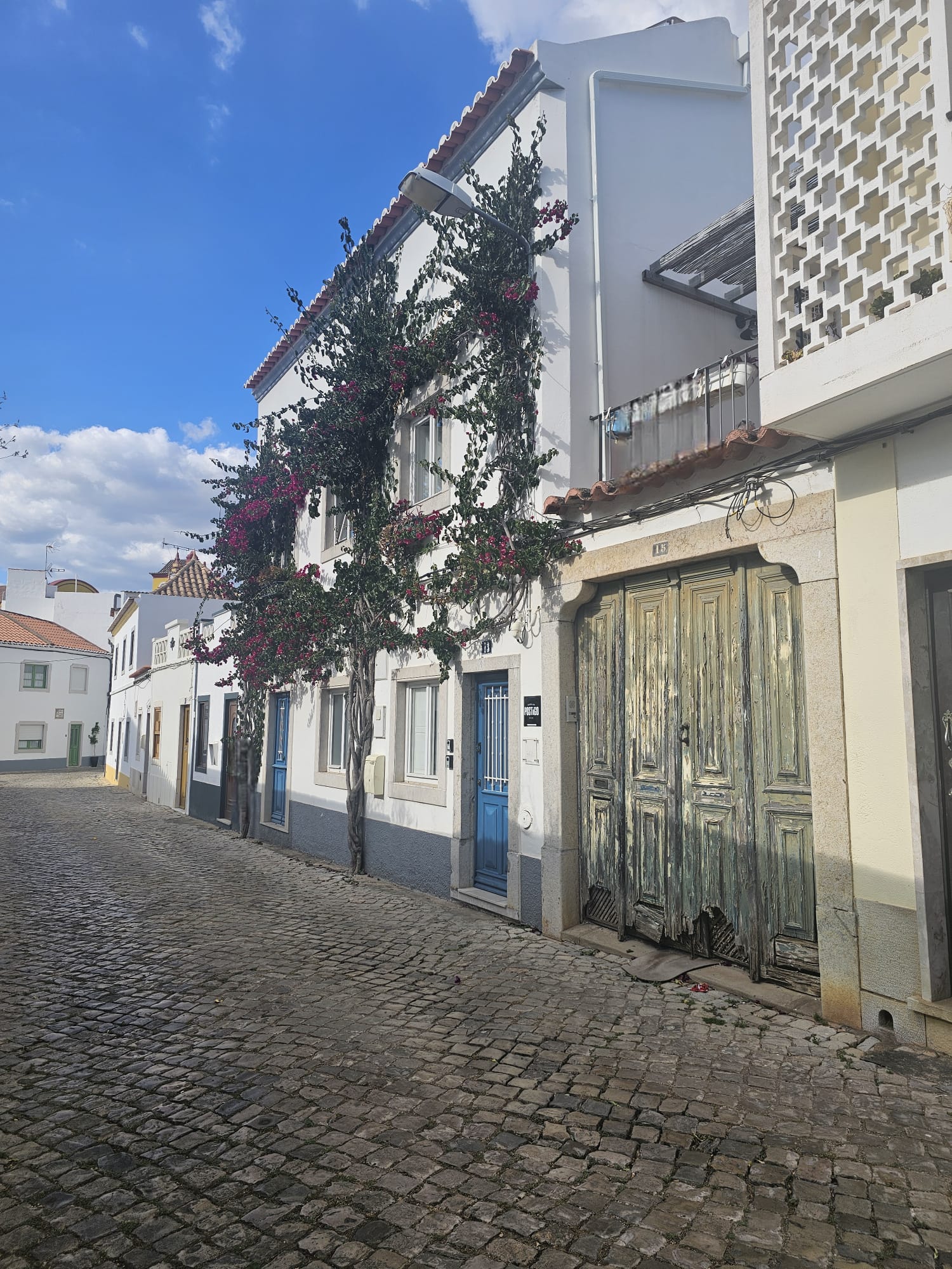 A charming cobblestone street lined with white buildings, one adorned with vibrant bougainvillea climbing its facade. The blue doors add a pop of color, while a weathered wooden door shows signs of age. The sky is bright blue, dotted with fluffy clouds.