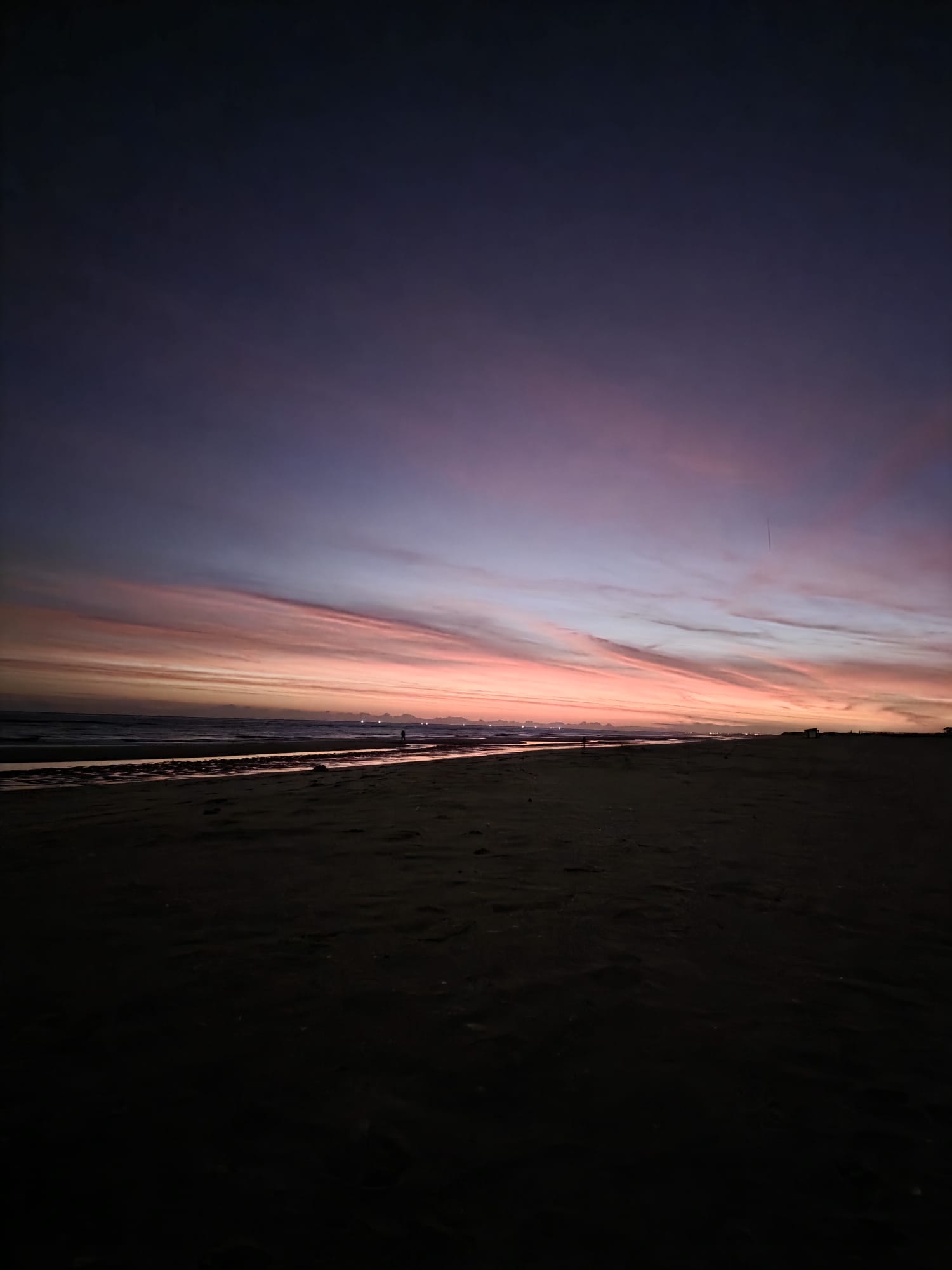  A wide-angle photo of a beach at sunset or sunrise, featuring a dramatic sky with vibrant streaks of pink, orange, and purple clouds above the dark sand and a wet shoreline