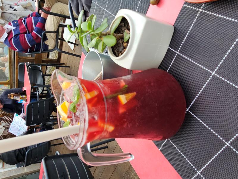A pitcher of red sangria filled with ice, orange slices, and mint sits on a checkered tablecloth next to a small potted plant on an outdoor café table. In the background, people are seated at other tables, enjoying their time.