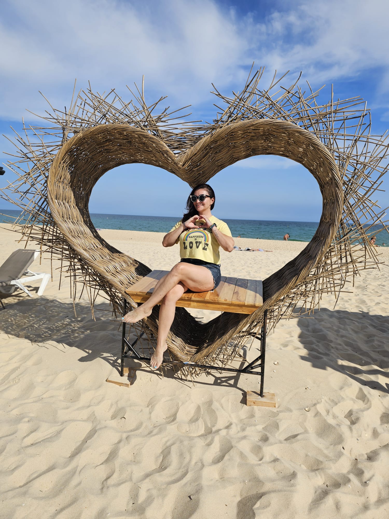 a woman sitting in a large heart shaped bench on the beach.