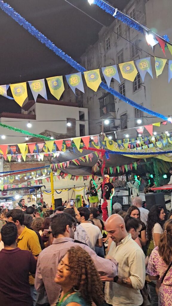 A lively Santos Populares street party in Lisbon with crowds dancing under colorful triangle bunting flags and streamers, with a live band performing on a small stage in the background