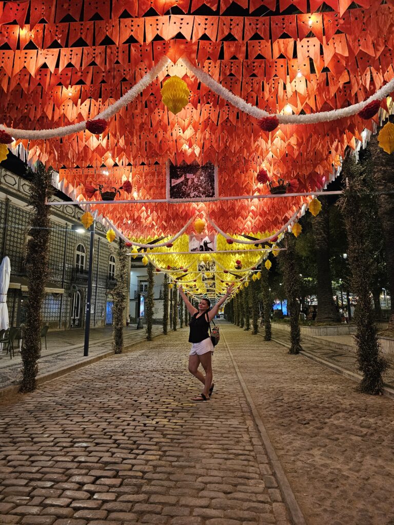A woman standing with her arms raised joyfully beneath a canopy of glowing red and yellow paper decorations lining a cobblestone street in Tavira at night during Santos Populares