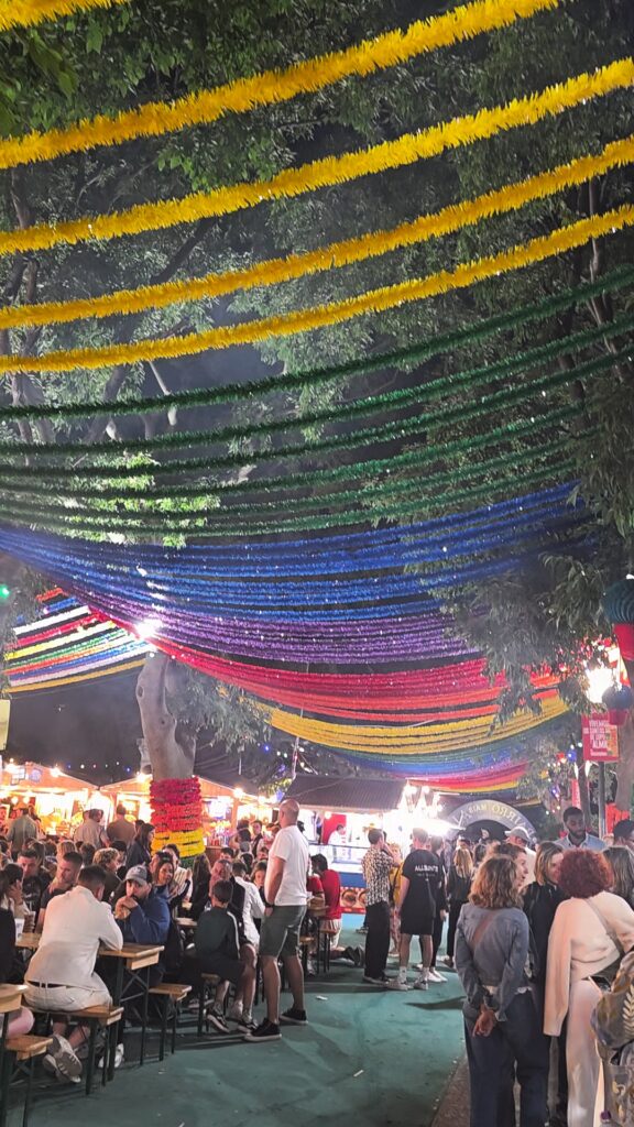 A packed Santos Populares street party in Lisbon at night with crowds of people under a canopy of brightly colored rainbow streamers hanging from the trees