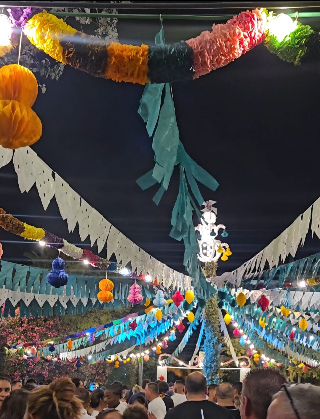 A crowd gathered at night beneath an elaborate canopy of colorful paper decorations, honeycomb lanterns, and bunting flags at a Santos Populares street party in Tavira, Portugal